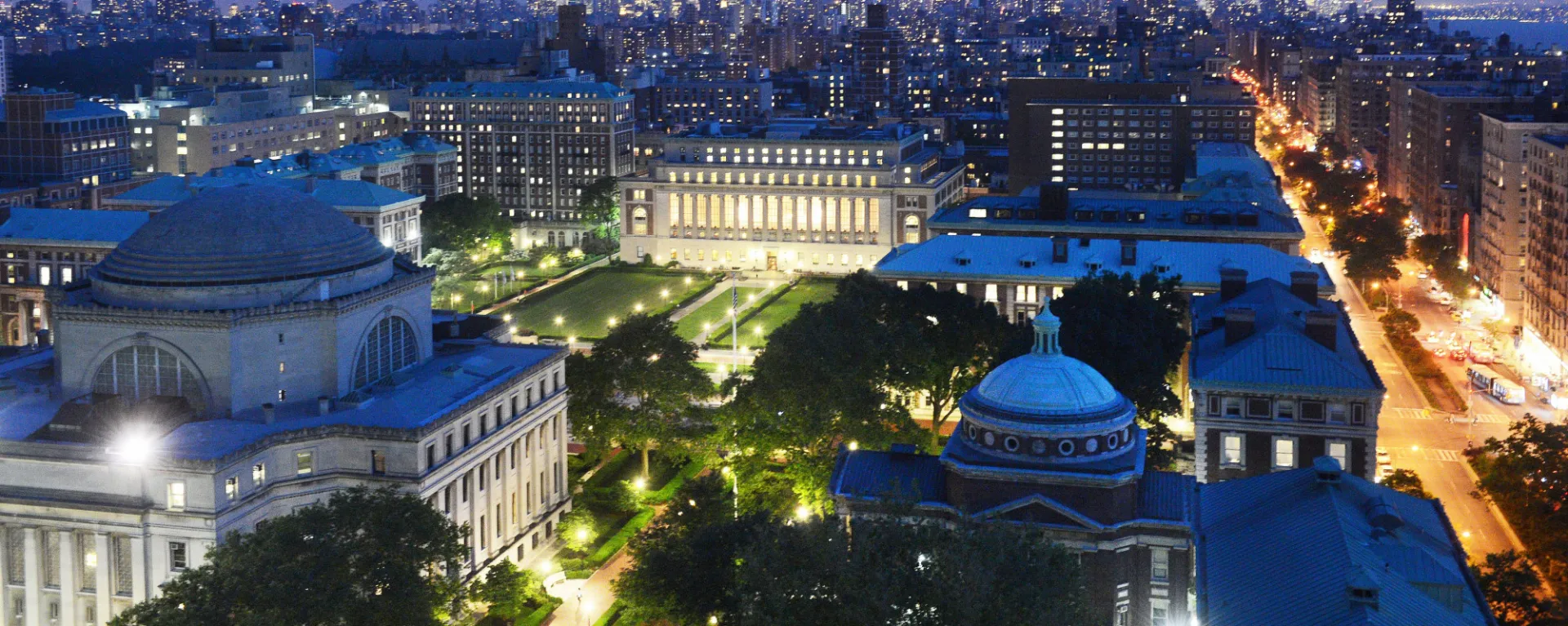 Aerial view of Columbia University’s campus at dusk with illuminated buildings and the Manhattan skyline
